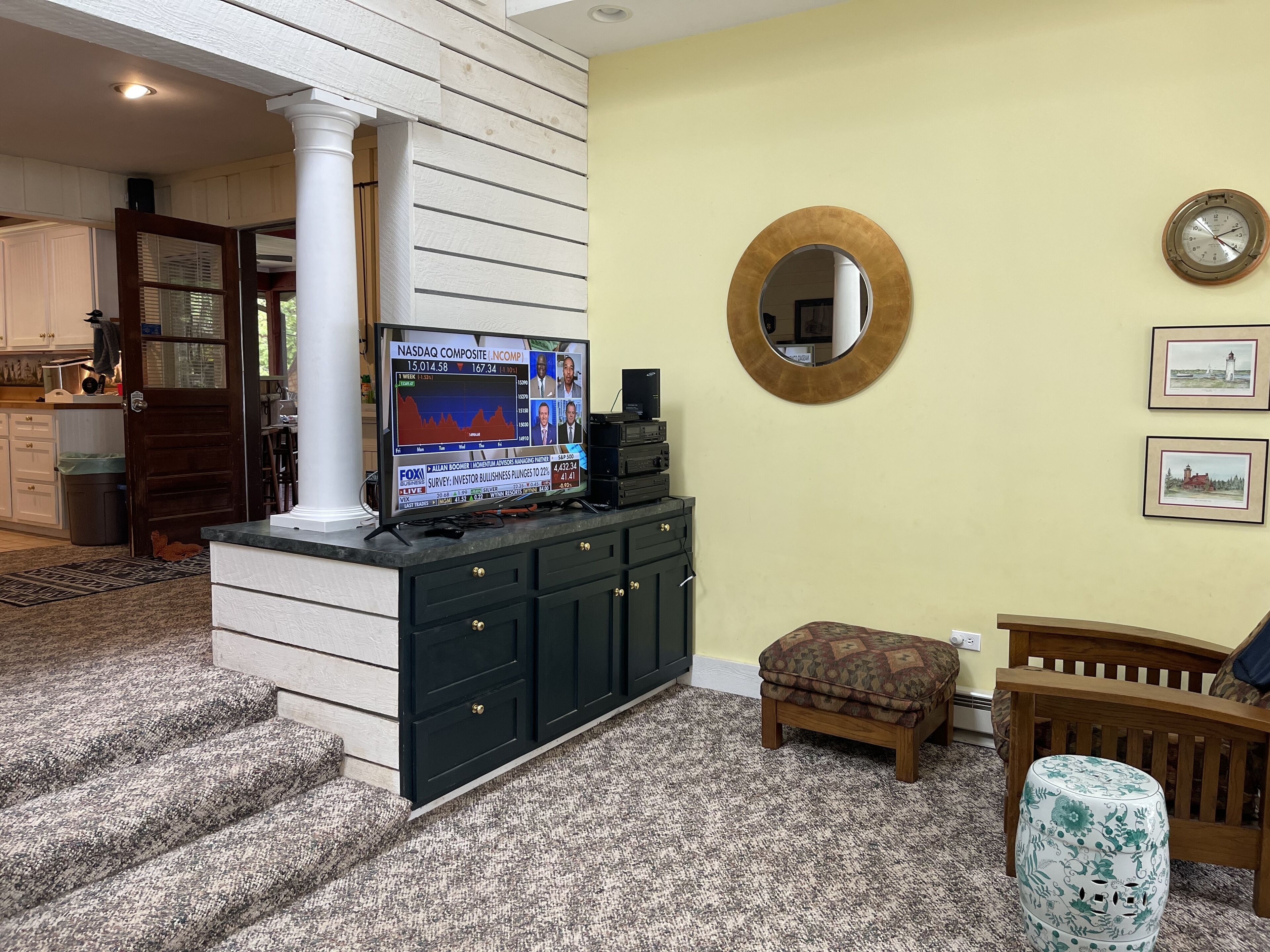 A living room with a television displaying financial news, a round mirror, and framed pictures on the wall.