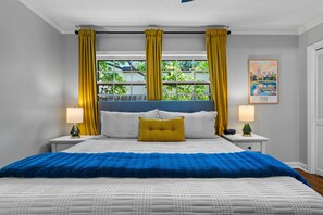 A bedroom with a bed, two bedside tables, a window with curtains, a cityscape painting, and a nightstand with a lamp. - EXPO's Cheerful 4-Bed w/Firepit Cherry St, UticaSq (Tulsa)