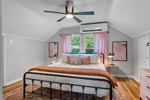 A bedroom with a bed, ceiling fan, air conditioning unit, windows with curtains, bedside lamps, and artwork on the walls.