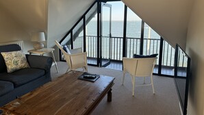 Een gezellige kamer met een houten bijzettafeltje, een bank en twee stoelen. Een groot raam biedt uitzicht op zee.