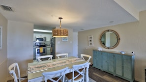 Uma sala de jantar com mesa de vidro, cadeiras brancas, luminária pendente de vime, armário verde-água e espelho redondo na parede.
