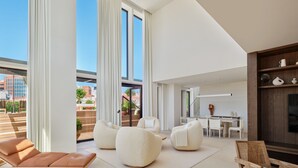 Ein modernes Wohnzimmer mit einer Couch, Sesseln und einem Holzstuhl. Es gibt einen Essbereich mit Tisch und Stühlen. Der Raum hat große Fenster und einen Blick auf die Stadt.