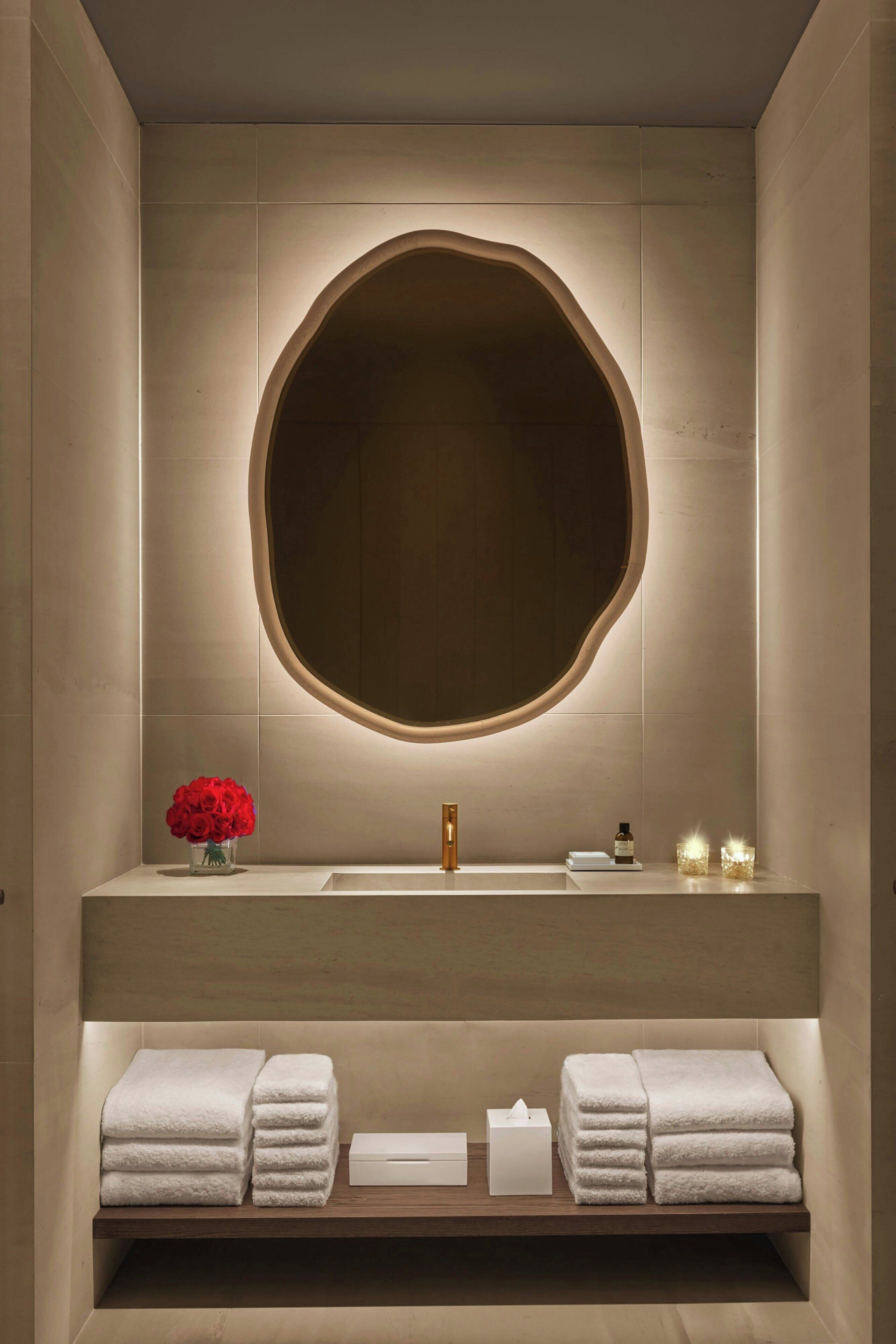 A modern bathroom with a large oval mirror, a sink, and a shelf with towels and toiletries.