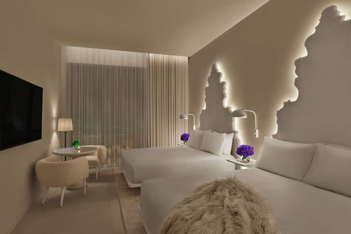 A modern hotel room with a large bed, a smaller bed, a round table with two chairs, a TV, and a decorative wall feature.
