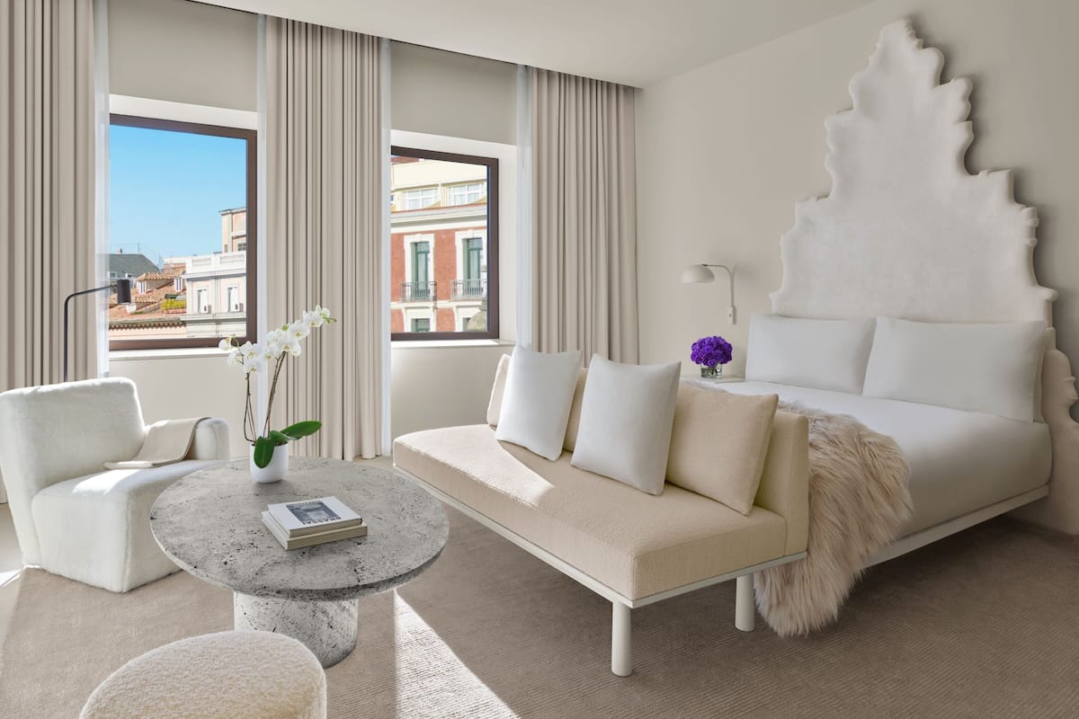 A modern hotel room with a white sofa, a round coffee table, and a view of the city through large windows.
