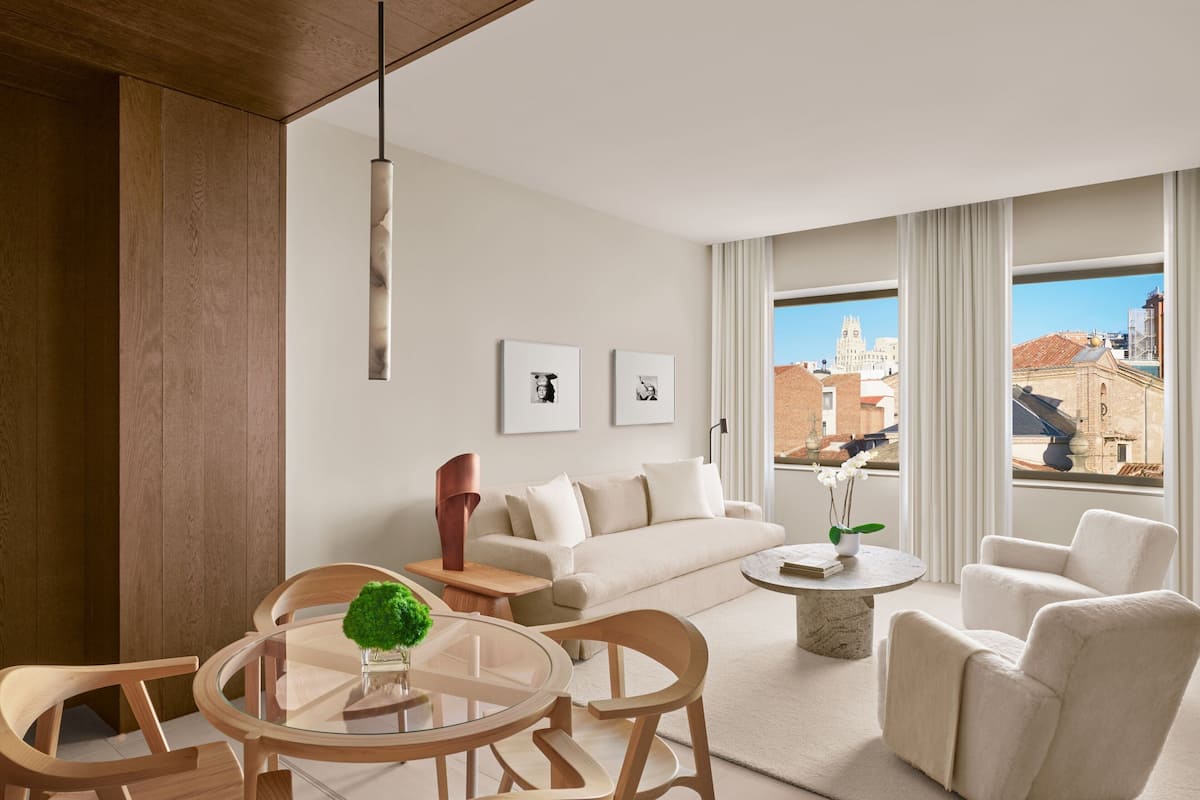 A modern living room with a wooden floor, a glass dining table, a beige sofa, and a view of buildings through large windows.