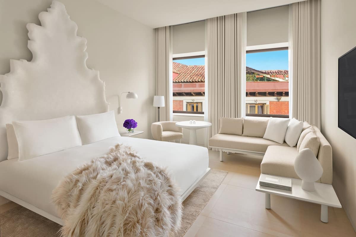 A modern hotel room with a large bed, a sofa, a small table, and a view of buildings through large windows.