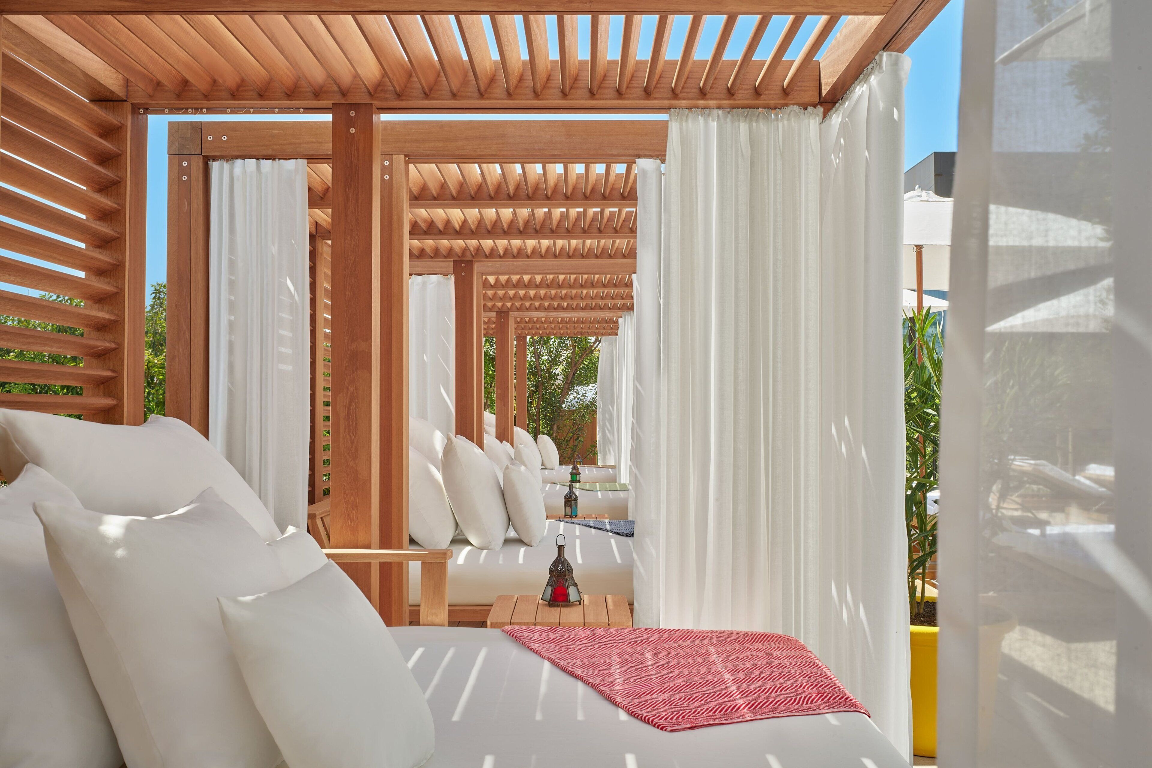 Un espace de détente extérieur moderne, agrémenté d’une pergola en bois, de coussins blancs et d’un tapis rouge tissé.