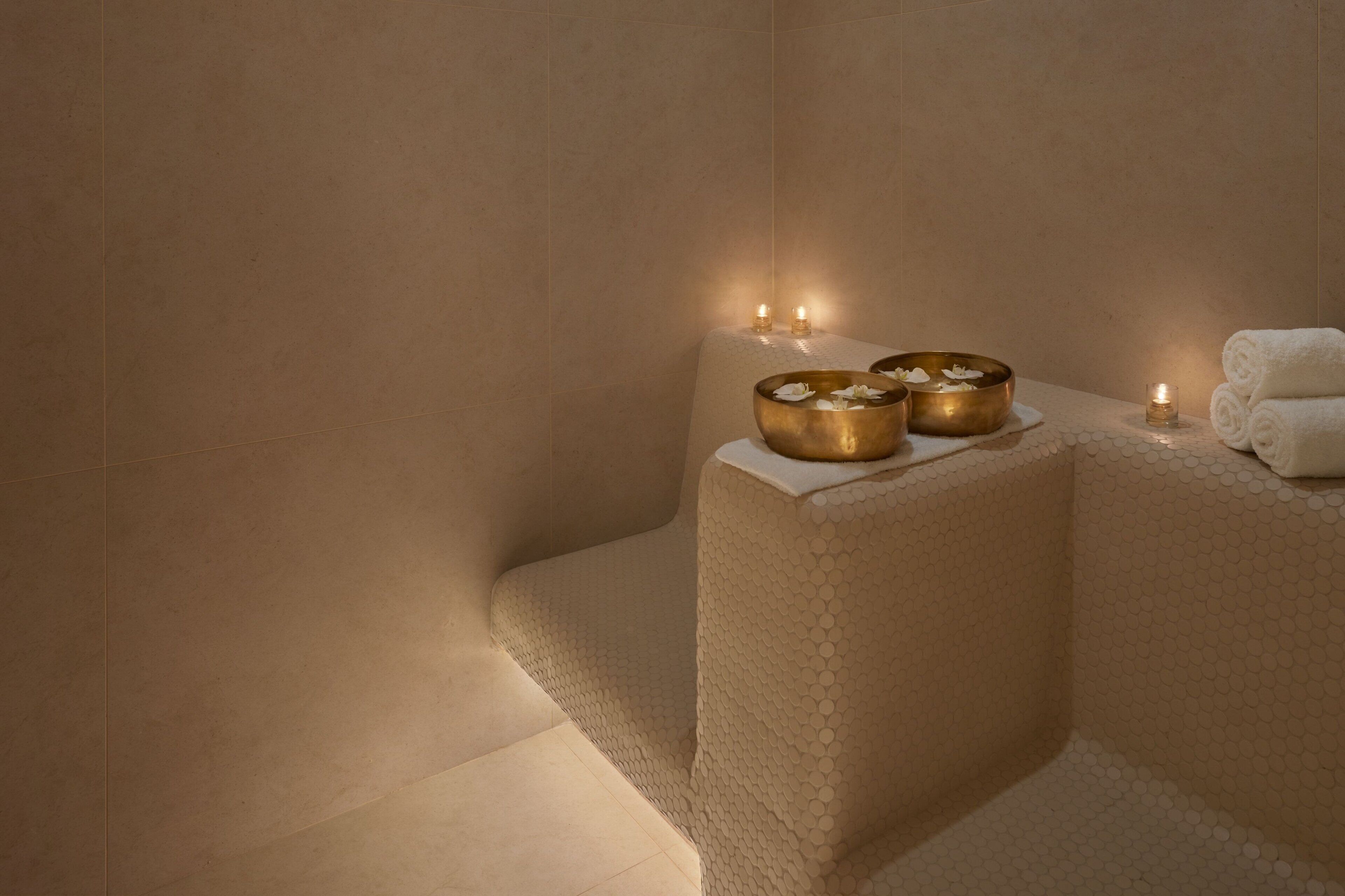 A spa area with a textured wall, two golden bowls on a white pedestal, and a rolled white towel on a white stand.