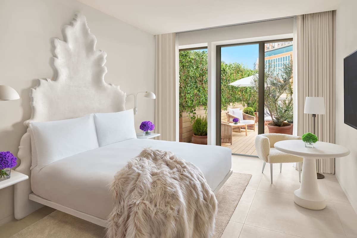 A modern hotel room with a large bed, a fluffy white rug, a round table with a lamp, and a view of the outdoor patio.