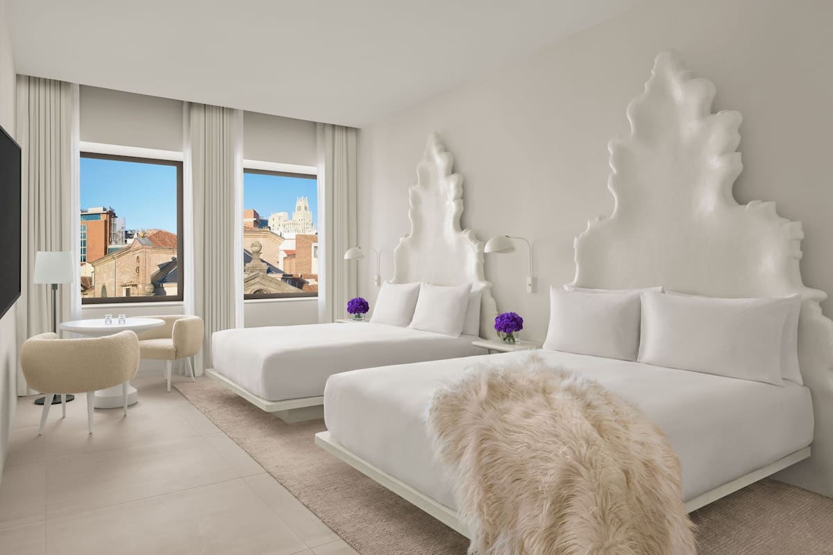A modern hotel room with two beds, a small sitting area, and a view of the cityscape.