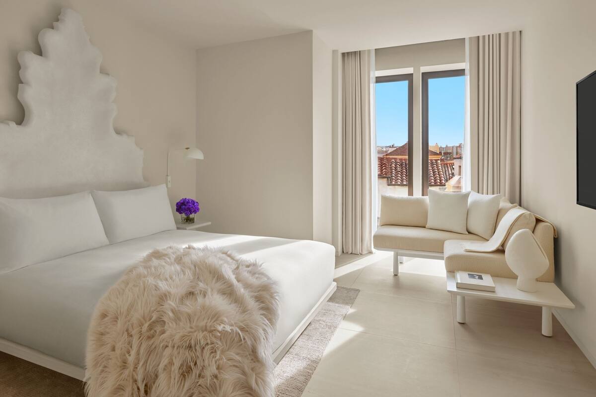 A modern hotel room with a large bed, a sofa, a coffee table, and a view of the cityscape through large windows.