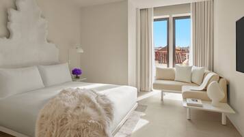 Quarto de hotel moderno com uma cama grande, um sofá, uma mesa de centro e vista para a paisagem urbana através de amplas janelas.