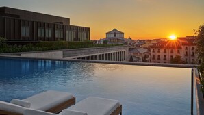 Ein Infinity-Pool auf dem Dach mit Liegestühlen, ein modernes Gebäude und im Hintergrund ein klassizistisches Gebäude bei Sonnenuntergang.