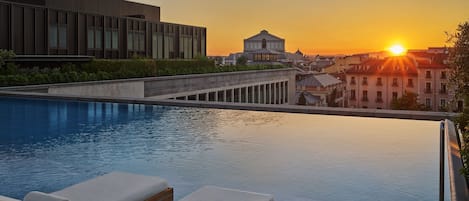 Una piscina infinita en la azotea con sillones de descanso, un edificio moderno y un edificio clásico al fondo durante el atardecer.