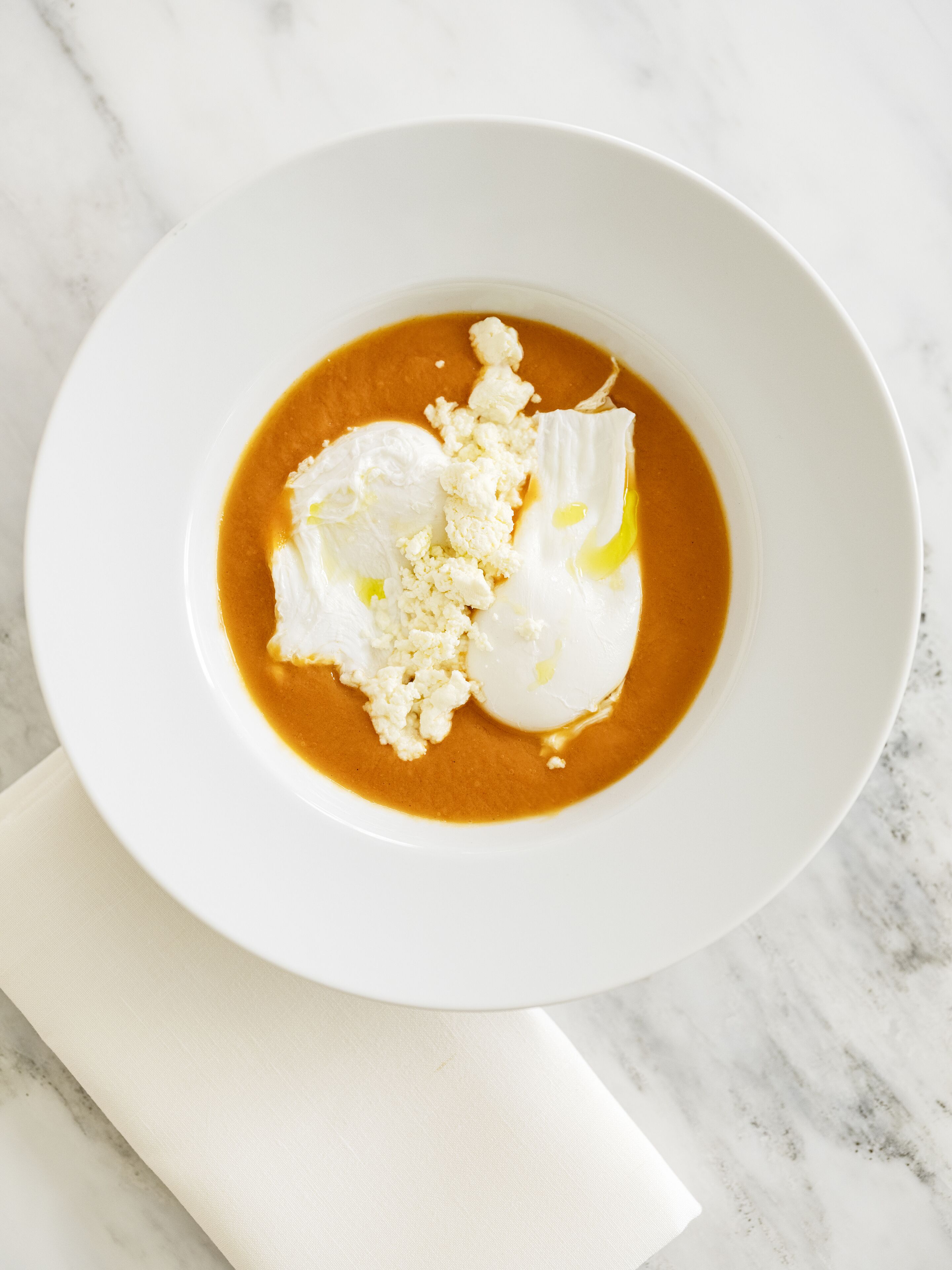 Un bol blanc contenant une soupe crémeuse, garni de fromage émietté et d’un œuf poché, posé sur une surface en marbre.