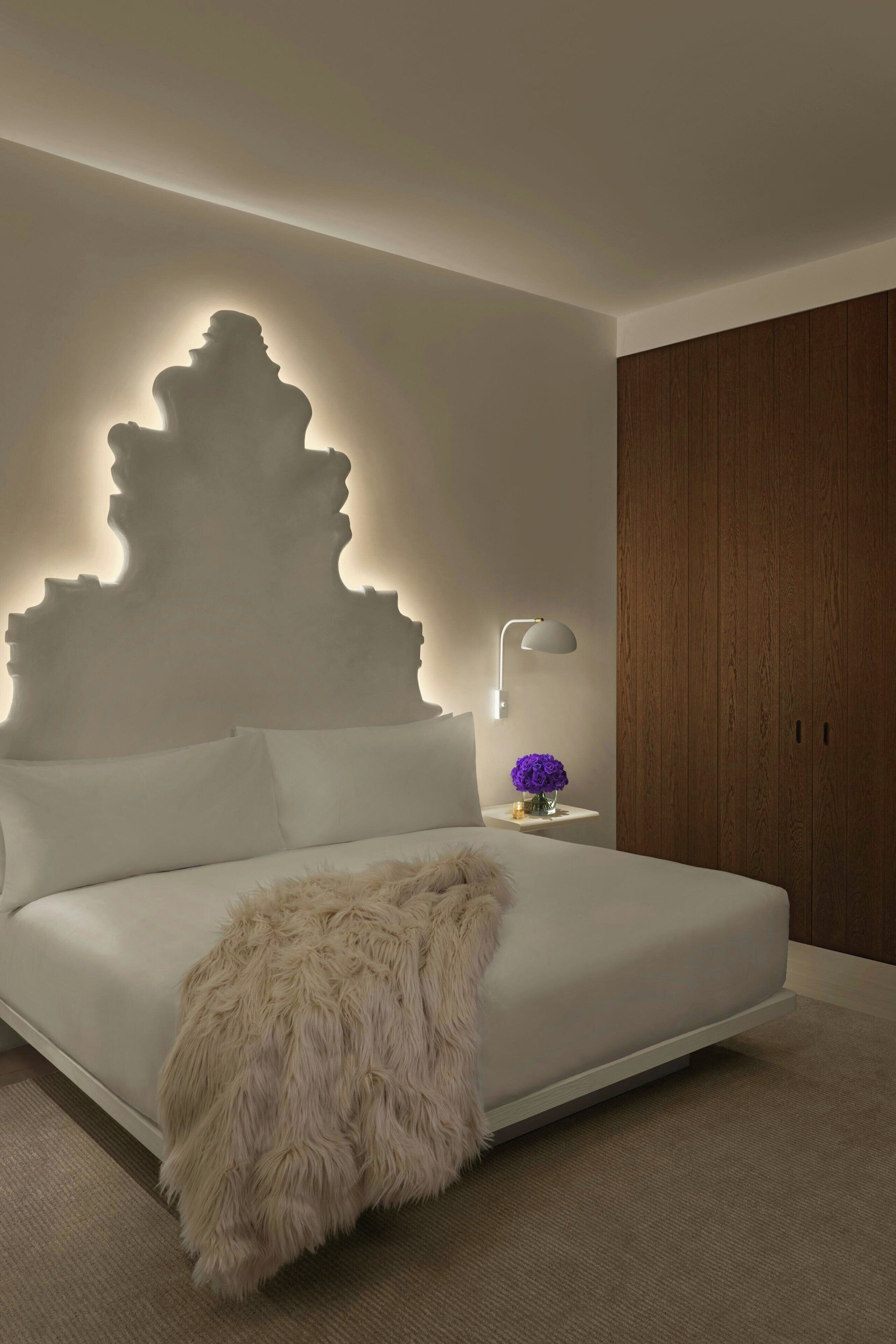 A modern bedroom with a white bed, a fluffy throw blanket, a bedside lamp, and a wall-mounted light fixture.