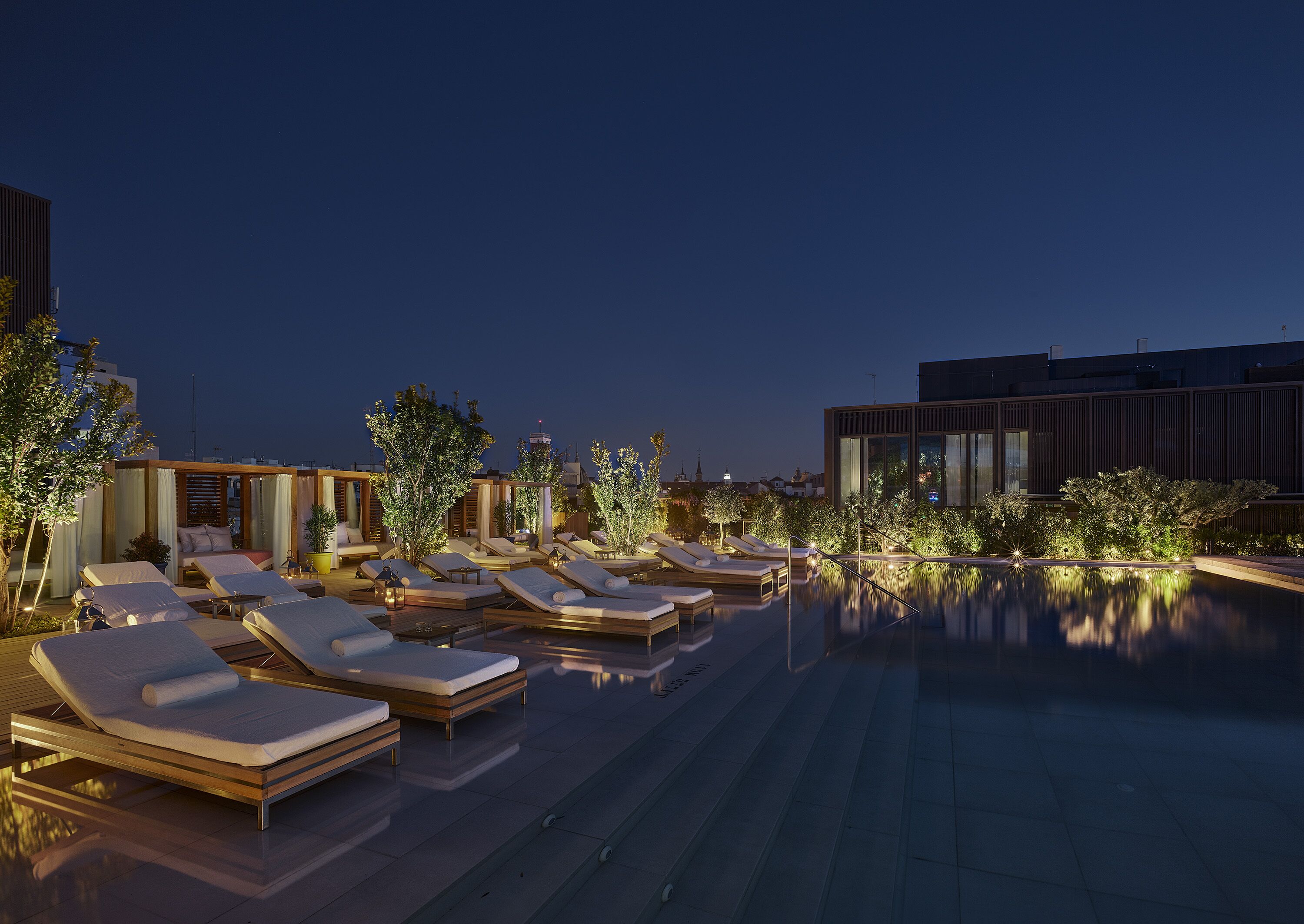 Un área de piscina en la azotea con tumbonas y un pequeño espacio de estar, rodeada de árboles y edificios modernos.