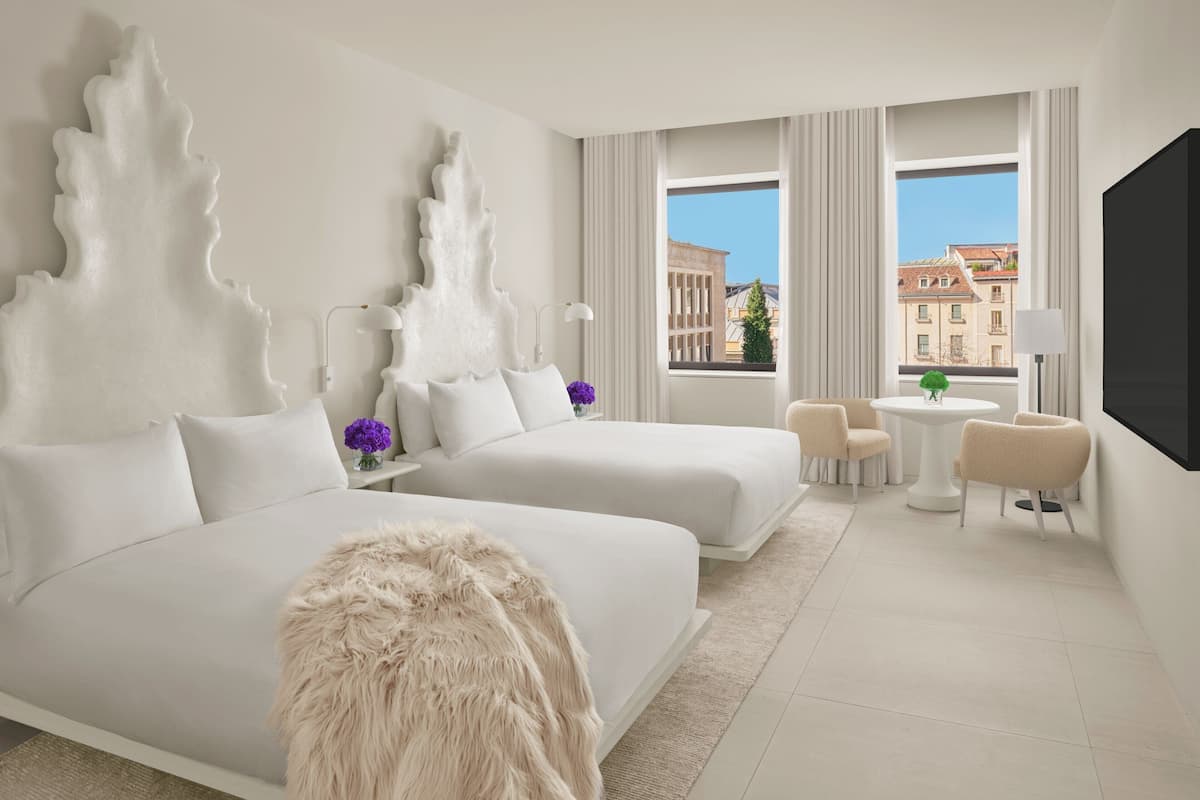 A hotel room with two beds, a large window, a flat-screen TV, a small round table, and a chair.