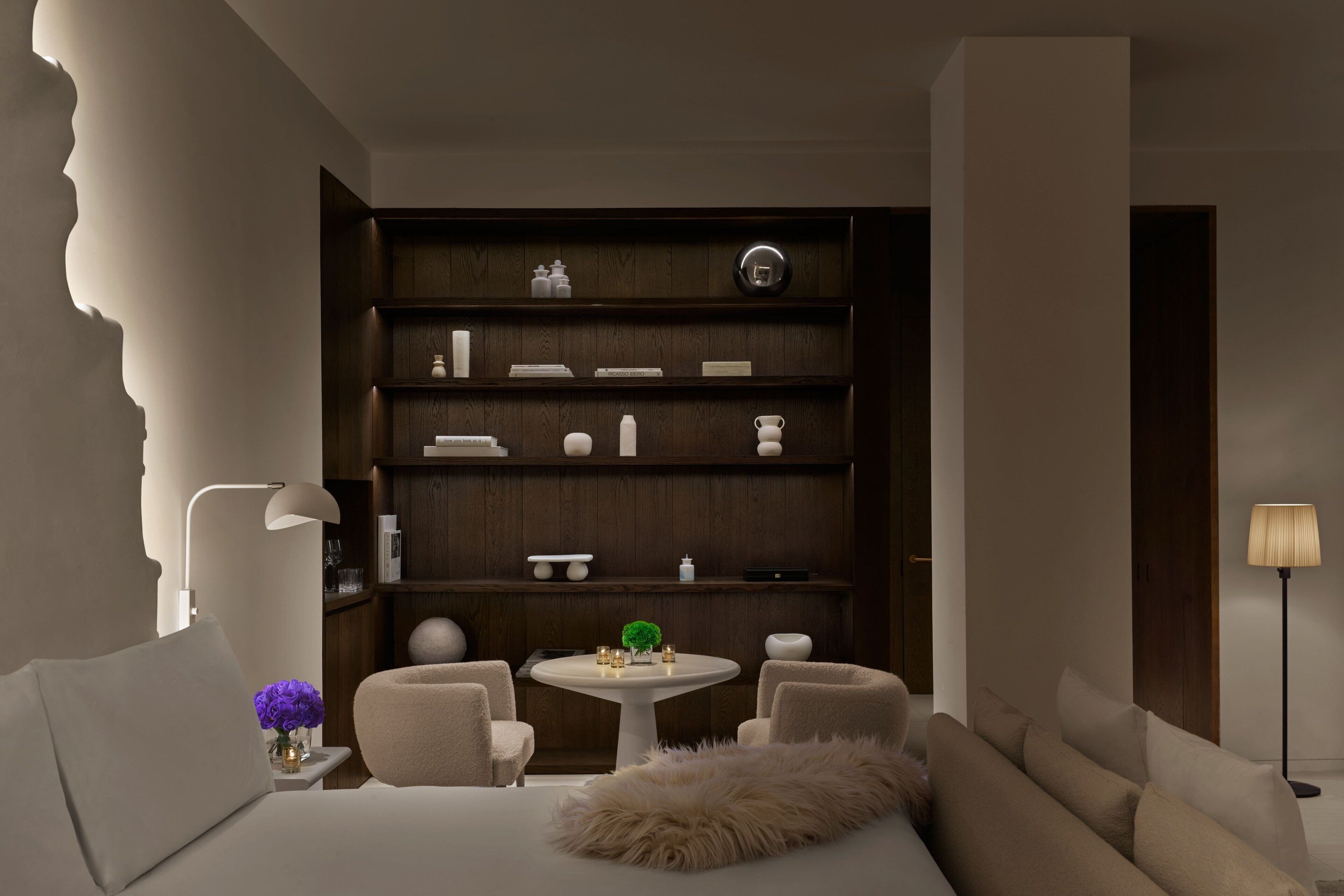 Habitación de hotel moderna con una cama grande, una mesa redonda con sillas, una estantería empotrada y un espejo amplio.