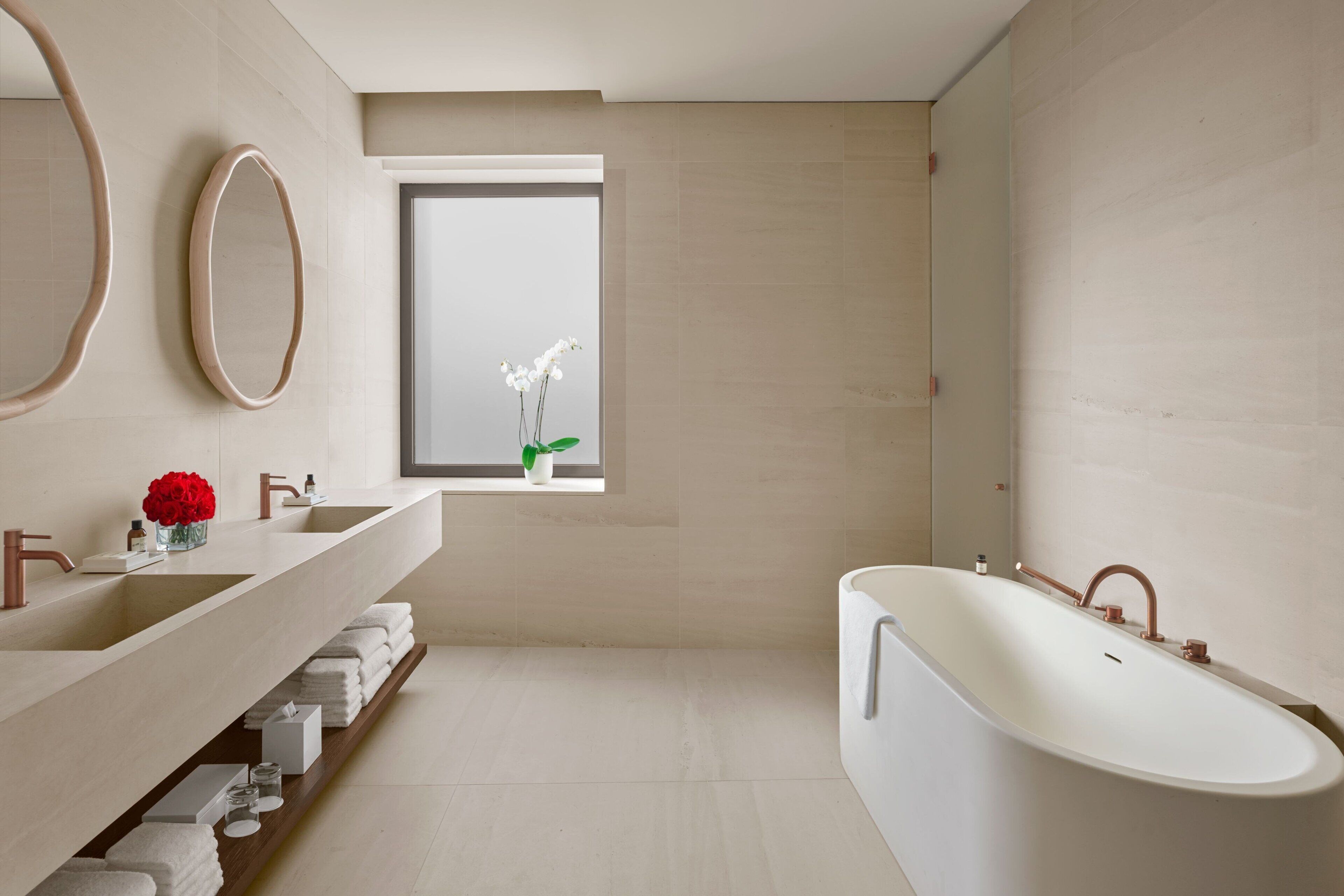 Une salle de bain moderne avec une grande baignoire, un meuble-lavabo avec un miroir et un vase de fleurs, et une fenêtre donnant sur l’extérieur.