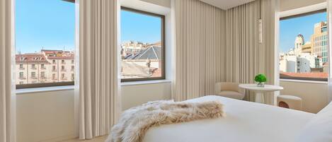 A hotel room with a large bed, a small round table, and a view of the cityscape through large windows.