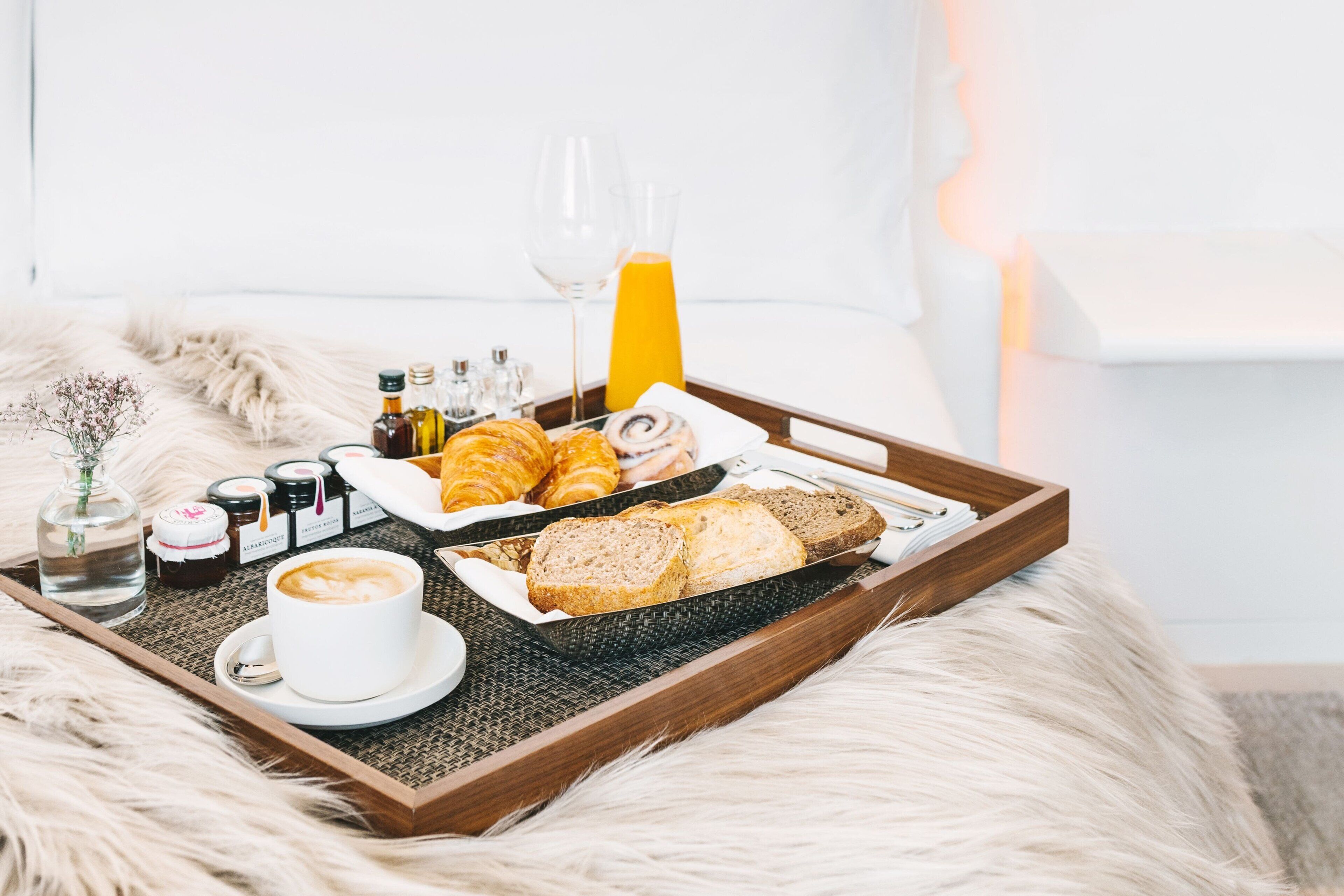 A breakfast tray with croissants, bread, coffee, and orange juice on a fluffy white bed.