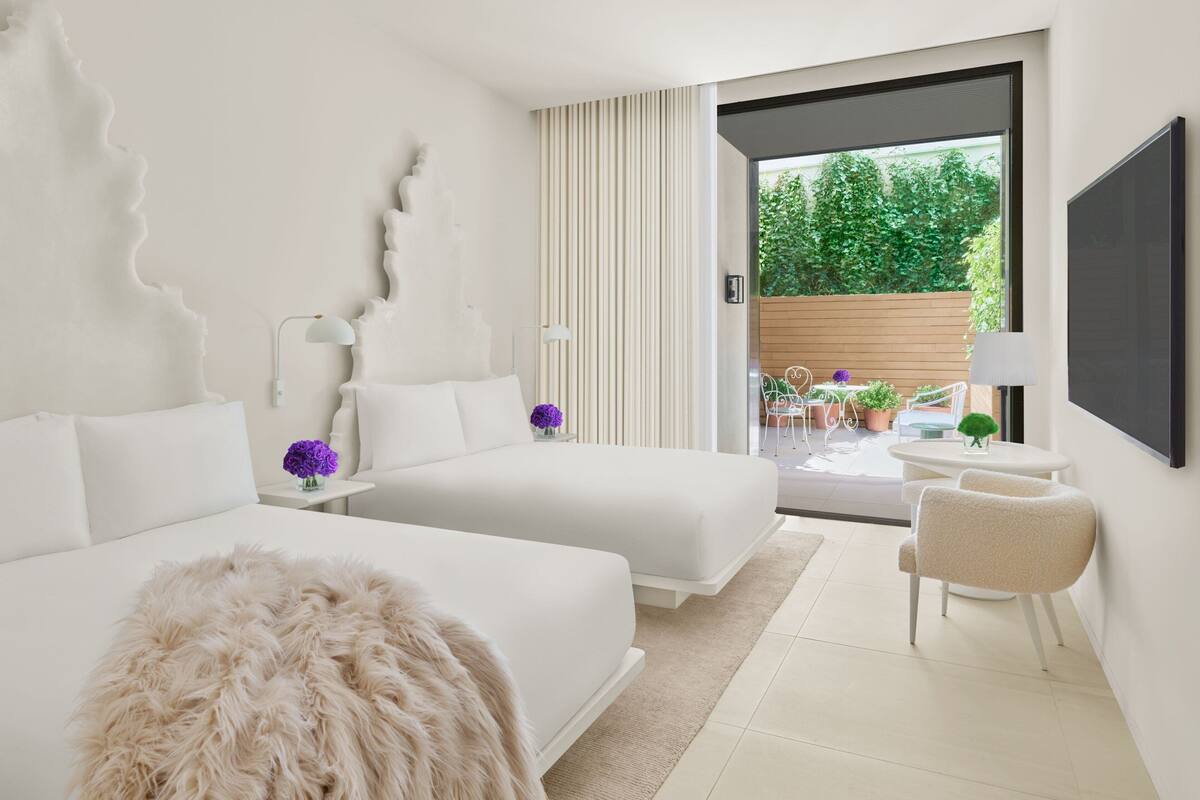 A modern hotel room with two beds, a large window, a flat-screen TV, and a balcony with plants.