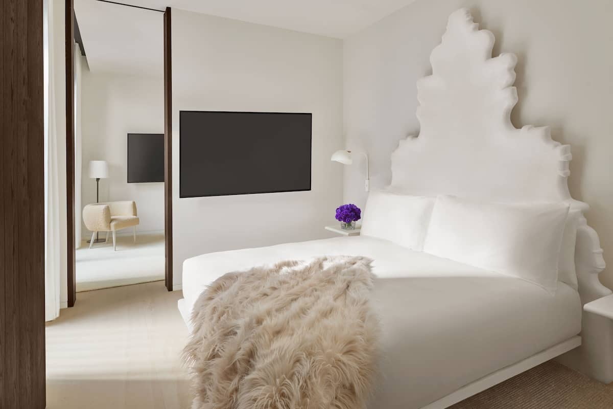 A modern bedroom with a large bed, a flat-screen TV, a white chair, and a decorative headboard.