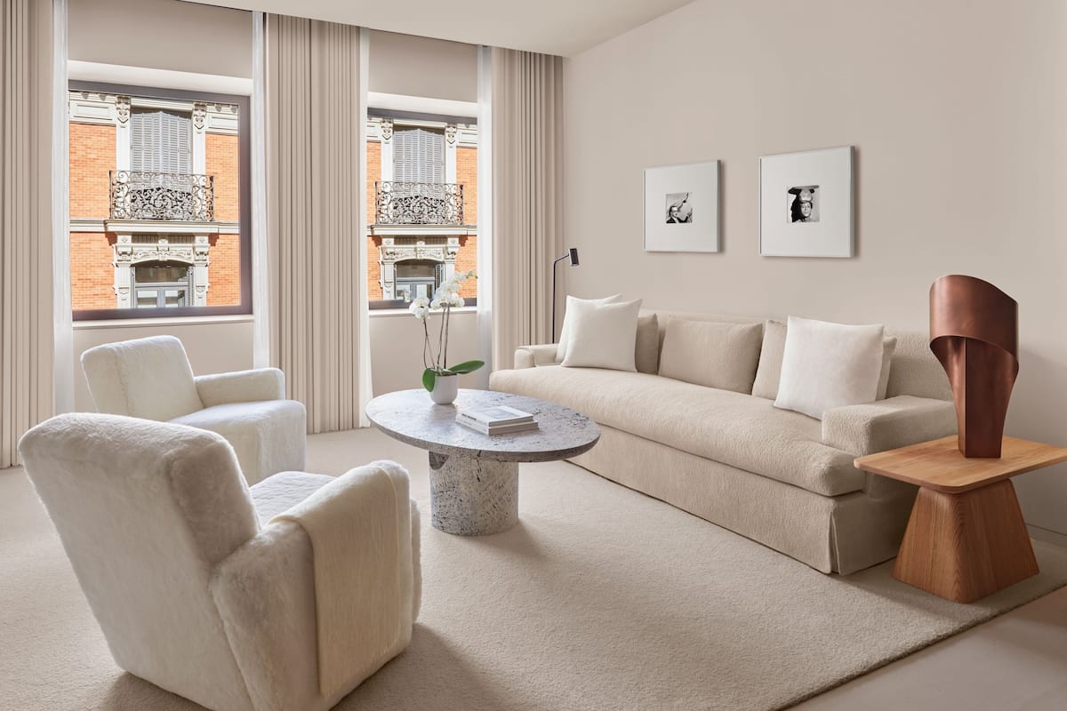 A modern living room with a beige sofa, white armchairs, a round coffee table, and a view of a brick building through large windows.