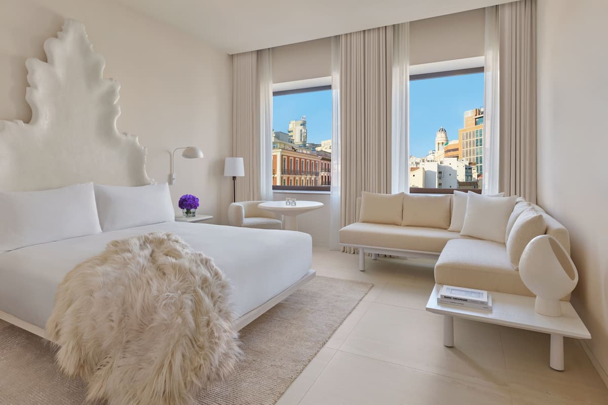 A modern hotel room with a large bed, a sofa, a small table, and a view of the city through large windows.