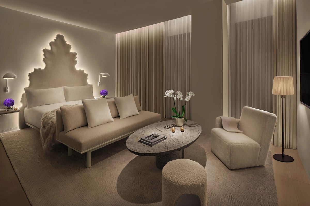 A modern hotel room with a sofa, a coffee table, a chair, and a bed with a unique headboard.