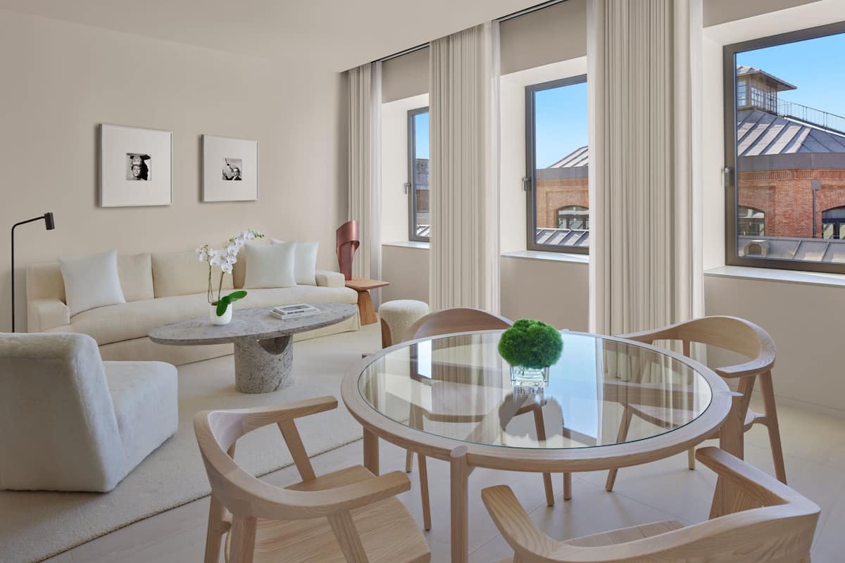A modern living room with a glass-top dining table, beige sofa, and a view of a brick building through large windows.