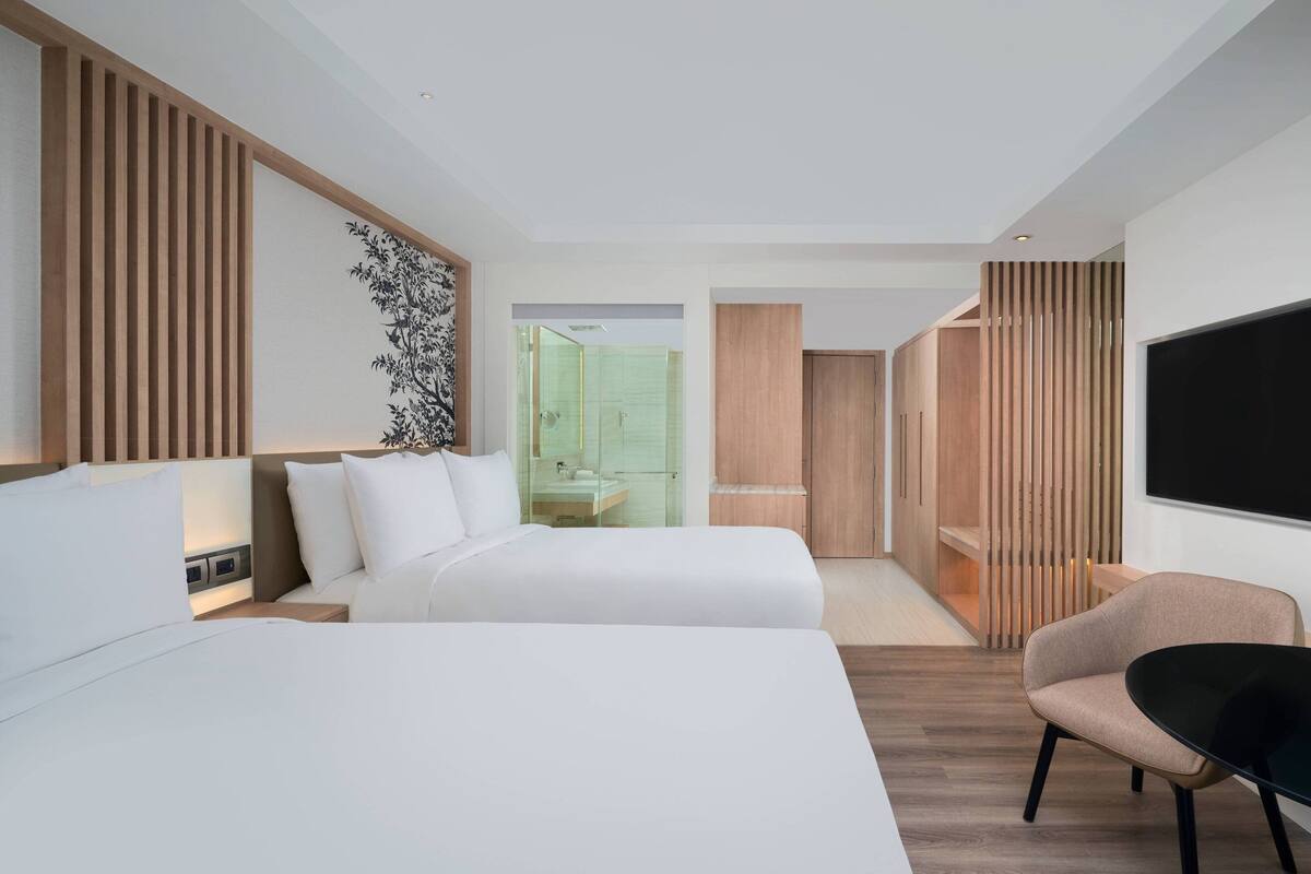 Une chambre d’hôtel moderne avec deux lits, un coin salon, une grande télévision et une salle de bain visible à travers une porte en verre.