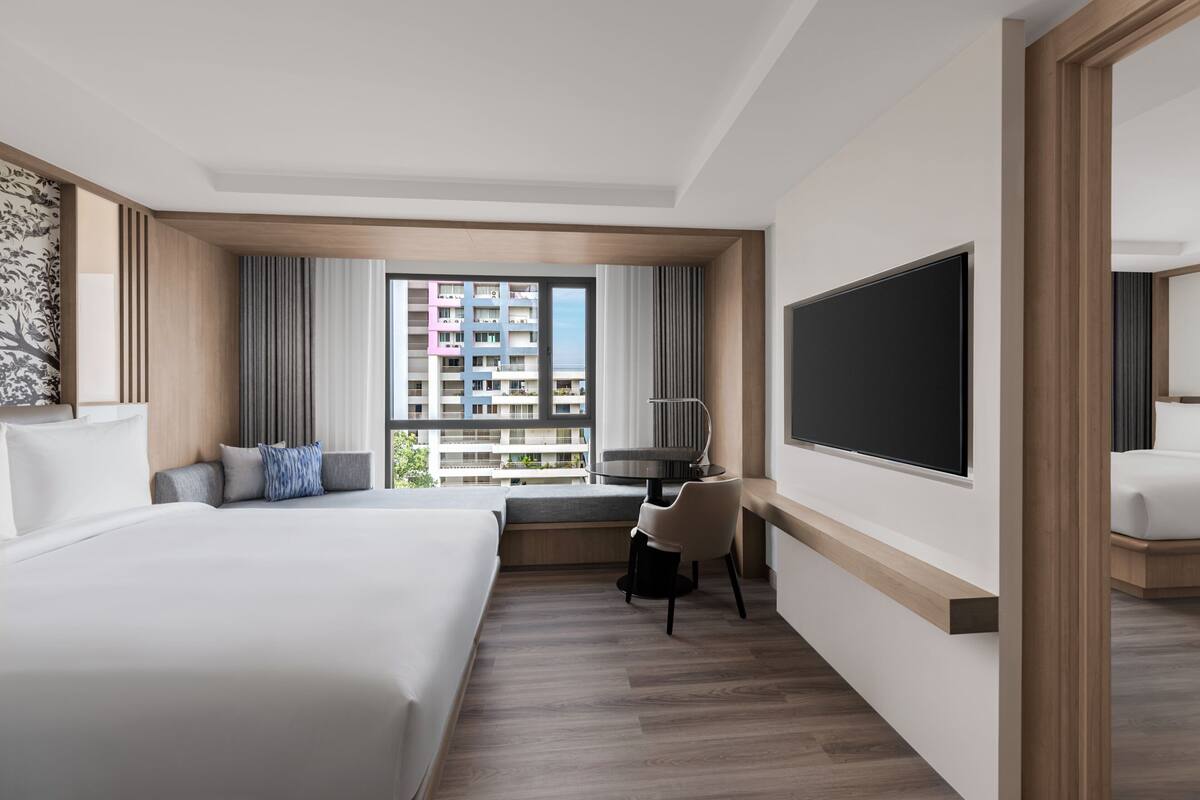 Une chambre d’hôtel moderne dotée d’un grand lit, d’un bureau avec une chaise, d’une grande fenêtre offrant une vue sur un immeuble de grande hauteur et d’un téléviseur à écran plat fixé au mur.