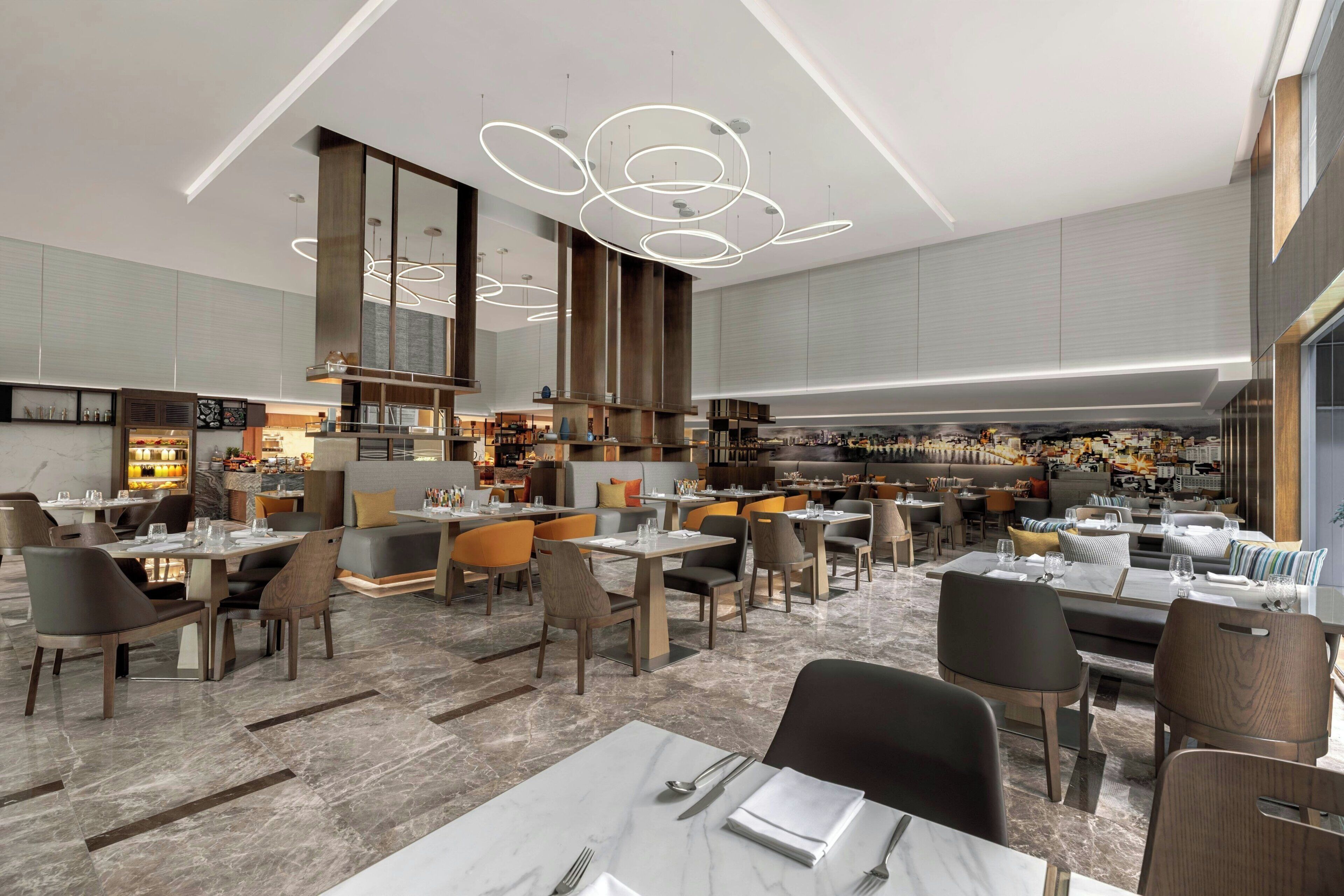 A modern restaurant with a central bar, multiple dining areas, and a high ceiling with circular light fixtures.