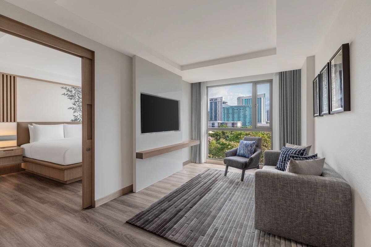 Une chambre d’hôtel moderne équipée d’un lit, d’un téléviseur fixé au mur, d’un canapé et offrant une vue sur le paysage urbain grâce à de grandes fenêtres.