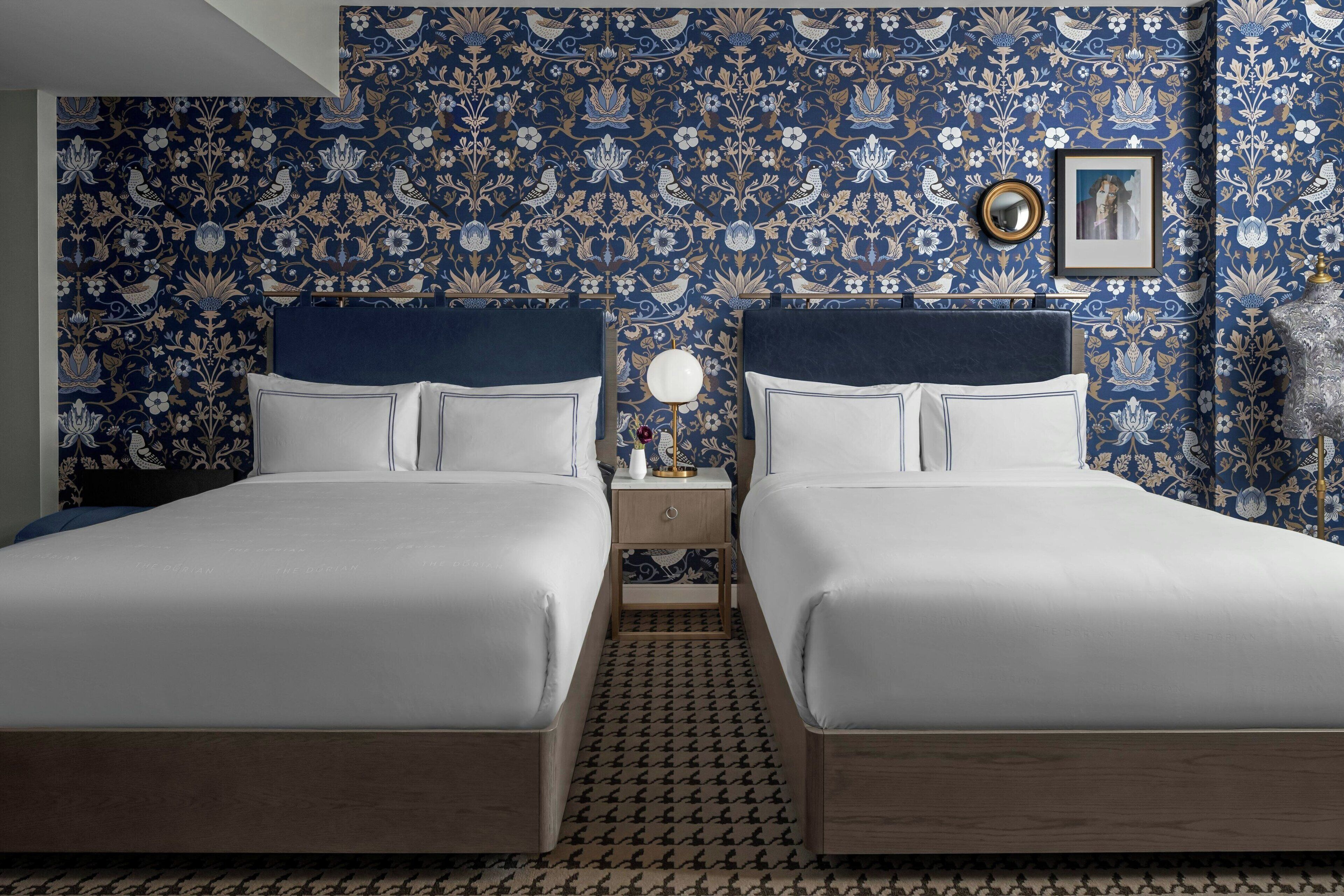 A double bed room with a patterned wallpaper, a bedside table with a lamp, and a framed picture on the wall.
