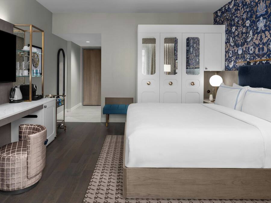 A modern hotel room with a large bed, a desk with a flat-screen TV, a seating area with a blue bench, and a patterned rug.