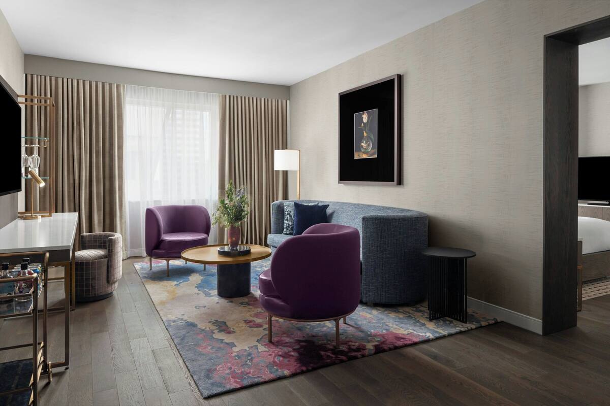 Una habitación de hotel moderna con sofá, dos sillones, una mesa de centro, televisor y un amplio ventanal con cortinas.
