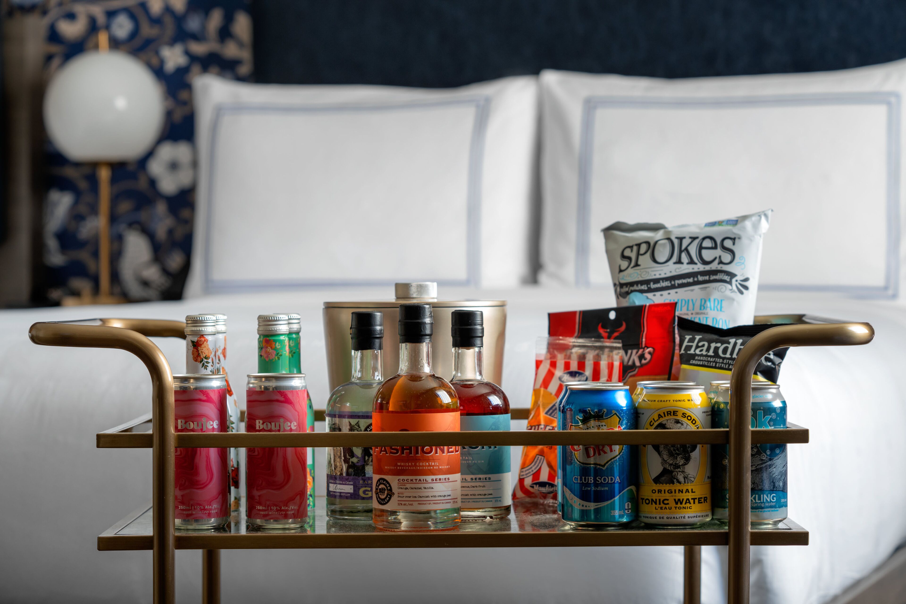 A minibar with various beverages and snacks, including bottles of alcohol, cans of soda, and a bag of chips, in a hotel room with a neatly made bed and decorative pillows.