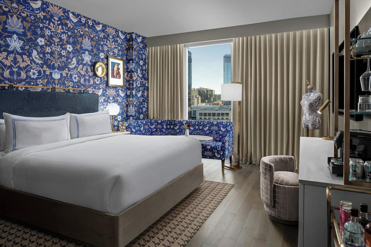 Habitación de hotel con una cama grande, un rincón de descanso, un tocador con espejo y vistas a la ciudad por la ventana.