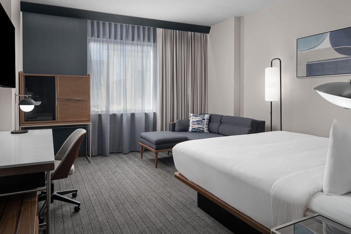 A hotel room with a bed, a desk, a chair, a sofa, a lamp, and a window with curtains.