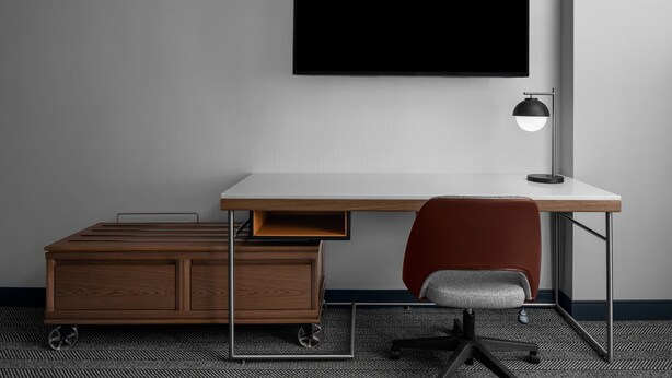 A modern office desk with a flat-screen monitor, a desk lamp, a red office chair, and a mobile cart with drawers. - Courtyard By Marriott Calgary Downtown (Calgary)