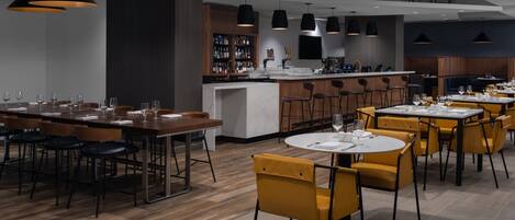 A modern restaurant with wooden flooring, yellow and black chairs, and a bar area with a wine display.