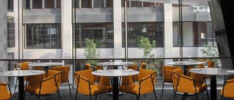 A modern dining area with round tables and orange chairs, large windows, and a view of the cityscape.