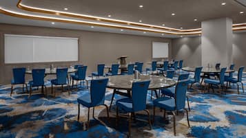 Un salón de conferencias con mesas redondas y sillas azules, suelo alfombrado y una pantalla de proyector.