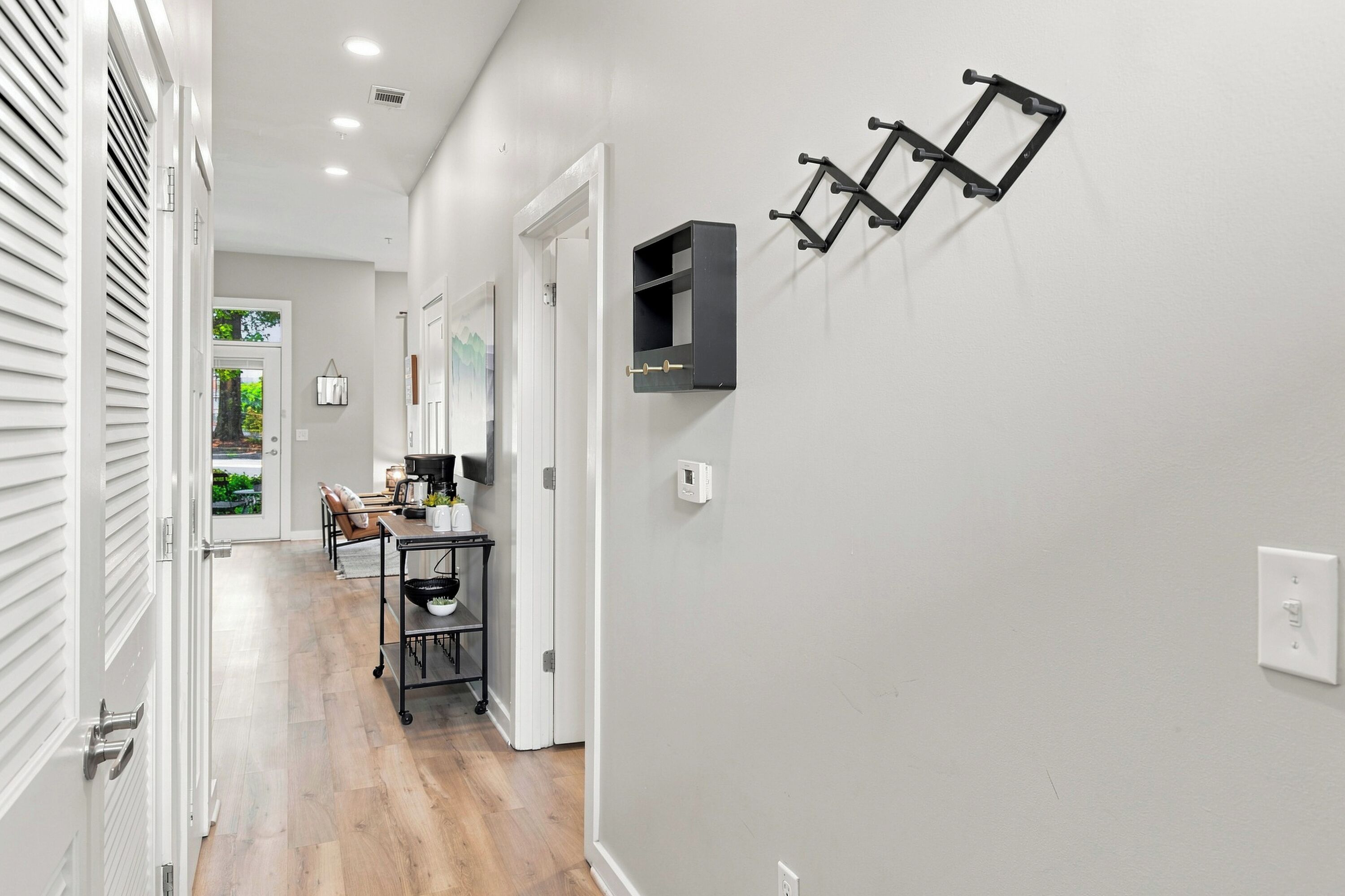 A hallway with a white door, a coat rack, a small table with a coffee maker, and a wall-mounted shelf.