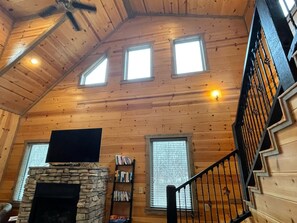 L’intérieur d’une cabane en rondins, avec une cheminée en pierre, un téléviseur à écran plat, une bibliothèque et un escalier doté d’une rampe en métal.