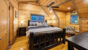 Dormitorio de cabaña de madera con cama, una silla, una mesa, ventilador de techo y ventana.